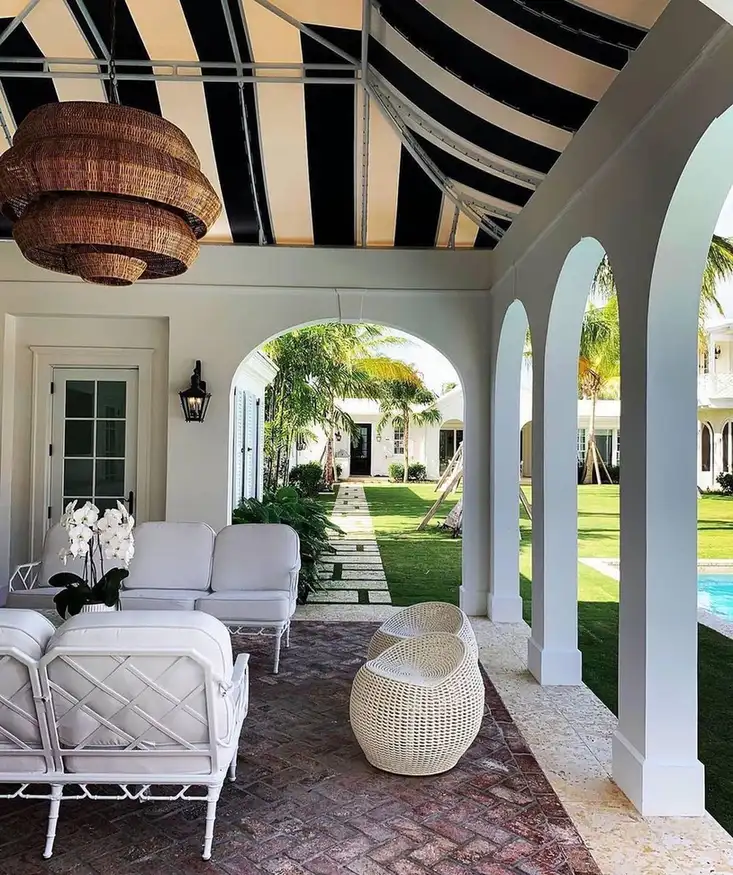 Screened covered porch with striped ceiling, pale seating, patterned rug, arched openings, and garden view