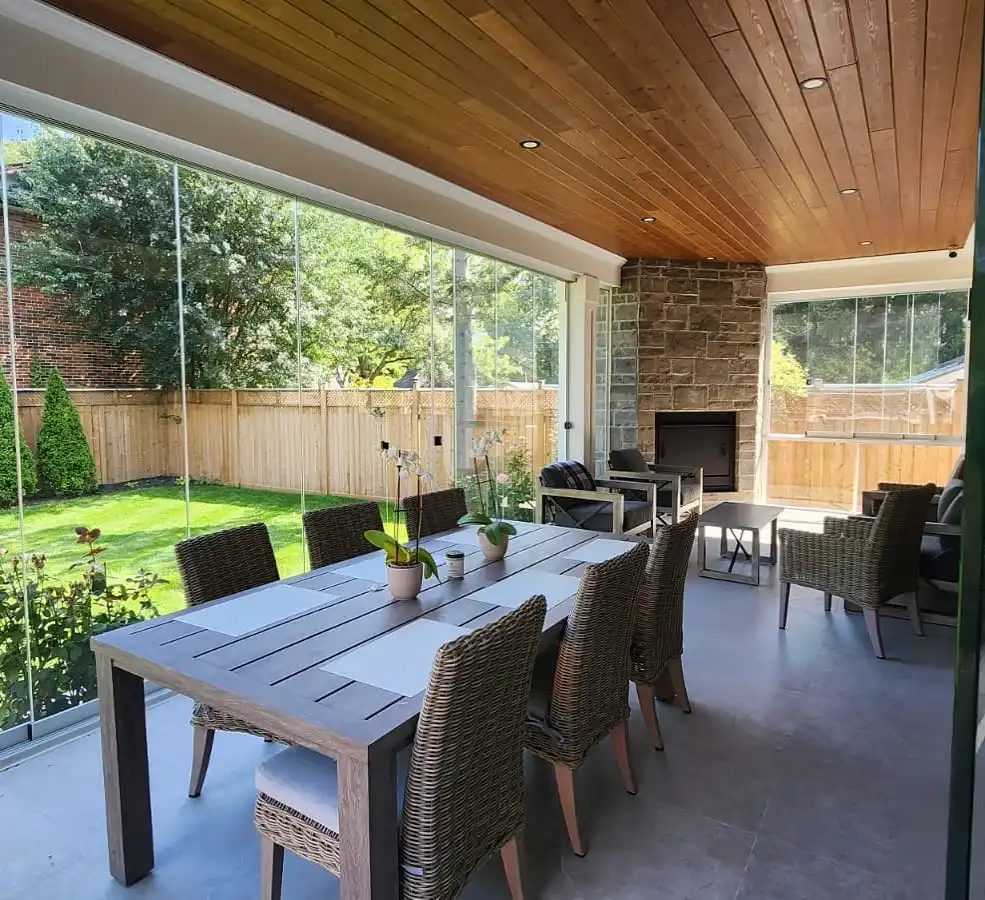 Glass-enclosed patio with floor-to-ceiling windows, wooden ceiling, dining table, woven chairs, and stone fireplace