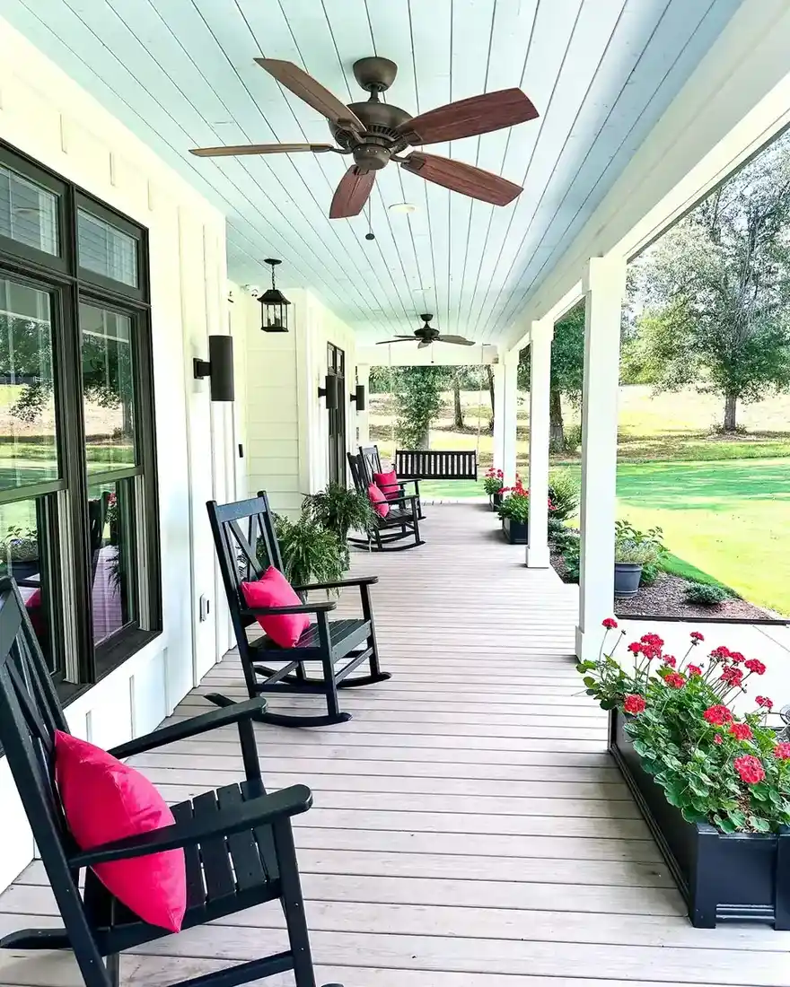 Long traditional covered porch with repeated rocking chairs, ceiling fans, flowers near the steps, and an open lawn.