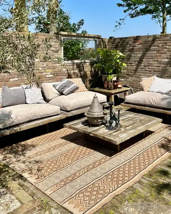 Layered natural outdoor rugs with earthy patterns beneath lounge seating, stone accents, and neutral cushions.
