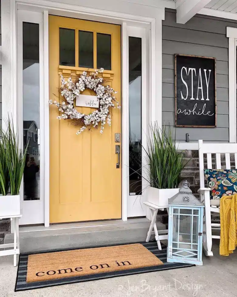 Yellow farmhouse front door with a wreath, black welcome sign, small bench, and white planter